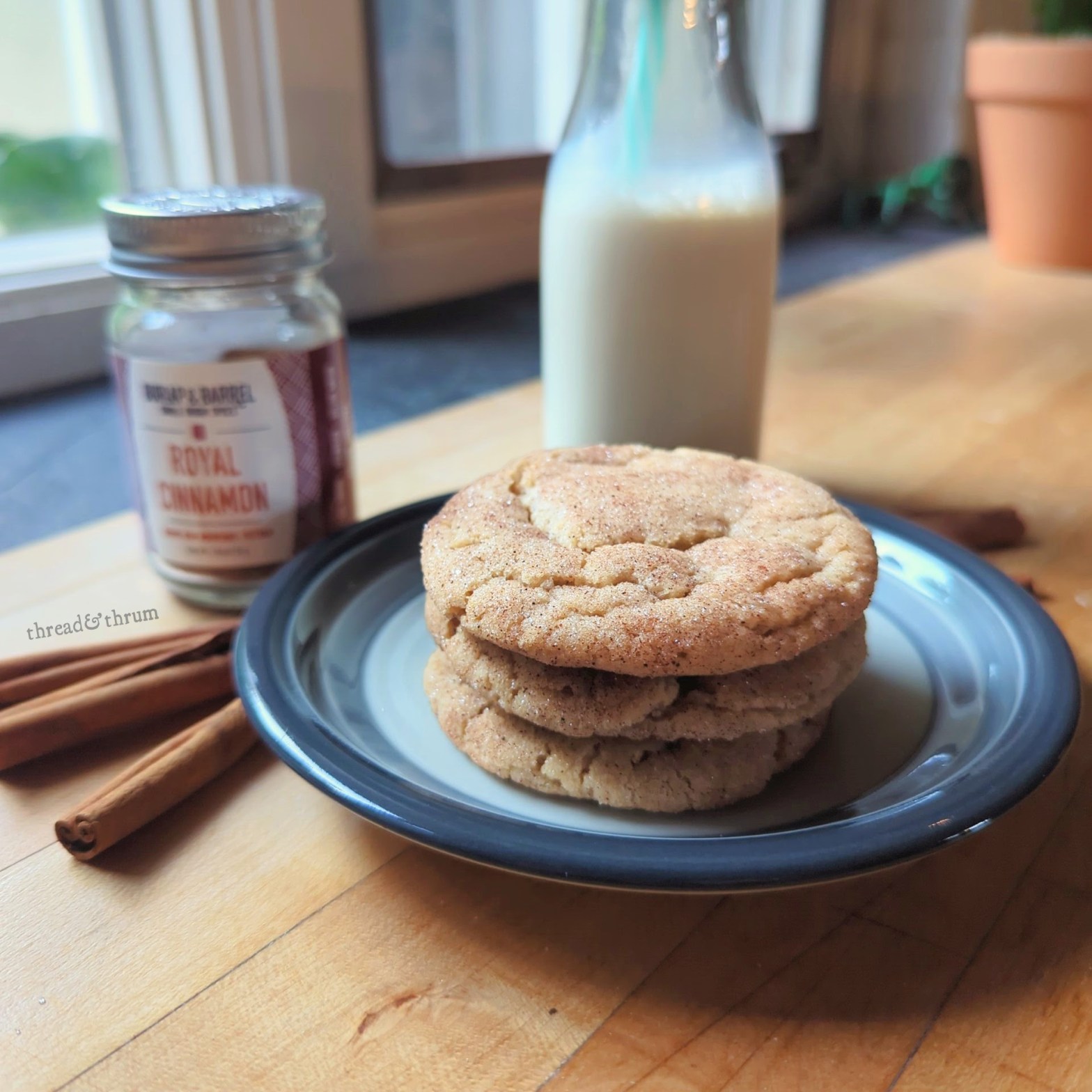 A stack of 3 snickerdoodles with cinnamon sugar coating, sitting on a blue and grey ceramic plate on a butcherblock counter. Surrounding the cookies are some cinnamon sticks, a jar of ground cinnamon, and a cute glass bottle of oat milk.