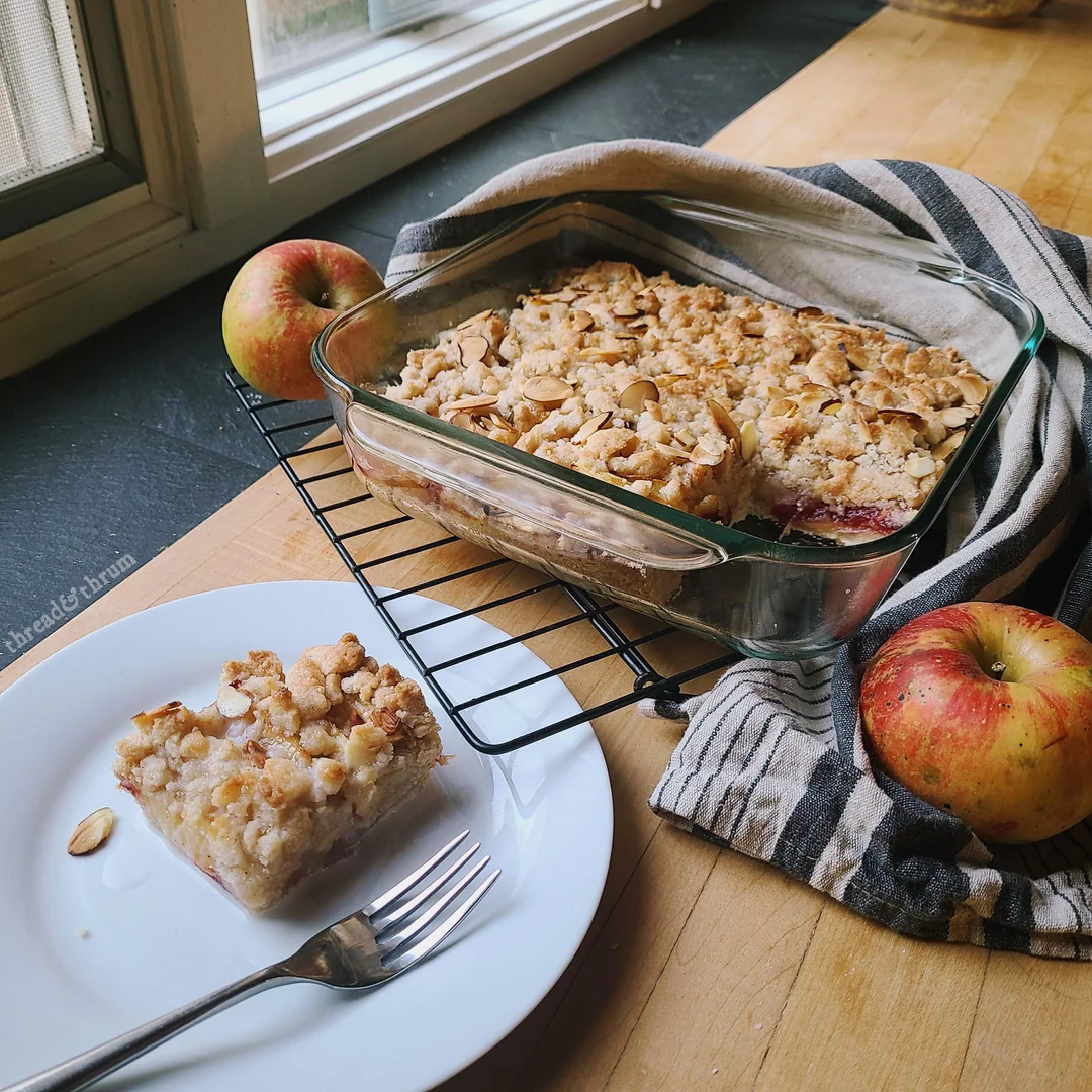 Angled view of a golden brown streusel topped cake in a square glass baking dish. A slice has been removed from the cake so its layers are visible: streusel topping, gooey fruit center, yeast cake base. It sits on a wooden butcher block and slate counter on a wire cooling rack, surrounded by a plate holding a slice of the cake, whole apples, and a blue and white IKEA hand towel