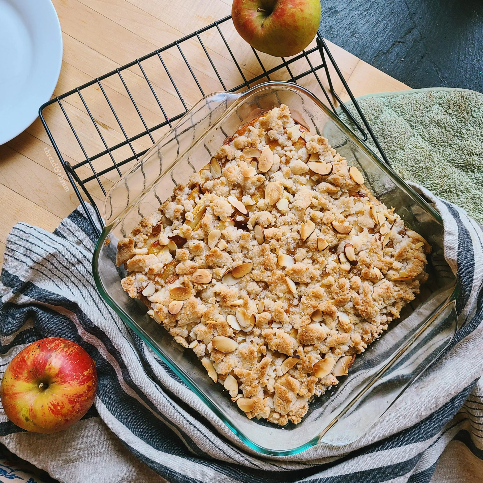 An overhead view of a golden brown, streusel-topped cake in a square glass cake pan. It sits on a cooling rack on a wooden counter, surrounded by a white and blue IKEA hand towel and a couple of whole Pink Lady apples