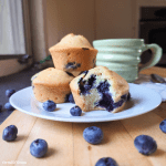 A plate of blueberry muffins, in which the front muffin has a bite taken out of it, showing a dense, regular crumb and blueberries. Behind this muffin is a golden mound of other muffins. Surrounding the plate of muffins are a scattering of blueberries and a handmade, ceramic, green glazed mug of coffee, all on a wooden countertop in a sunny kitchen.