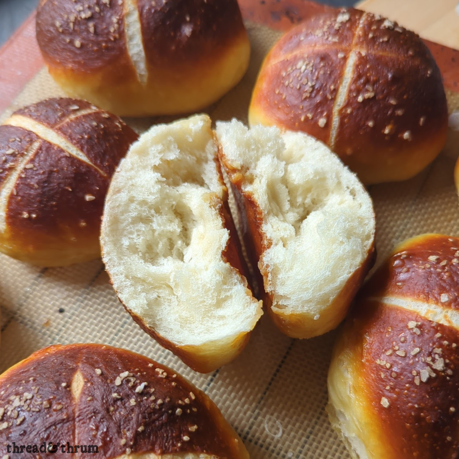 A closeup shot of a soft pretzel roll torn in half to show the pillowy soft interior. It's surrounded by whole pretzel rolls