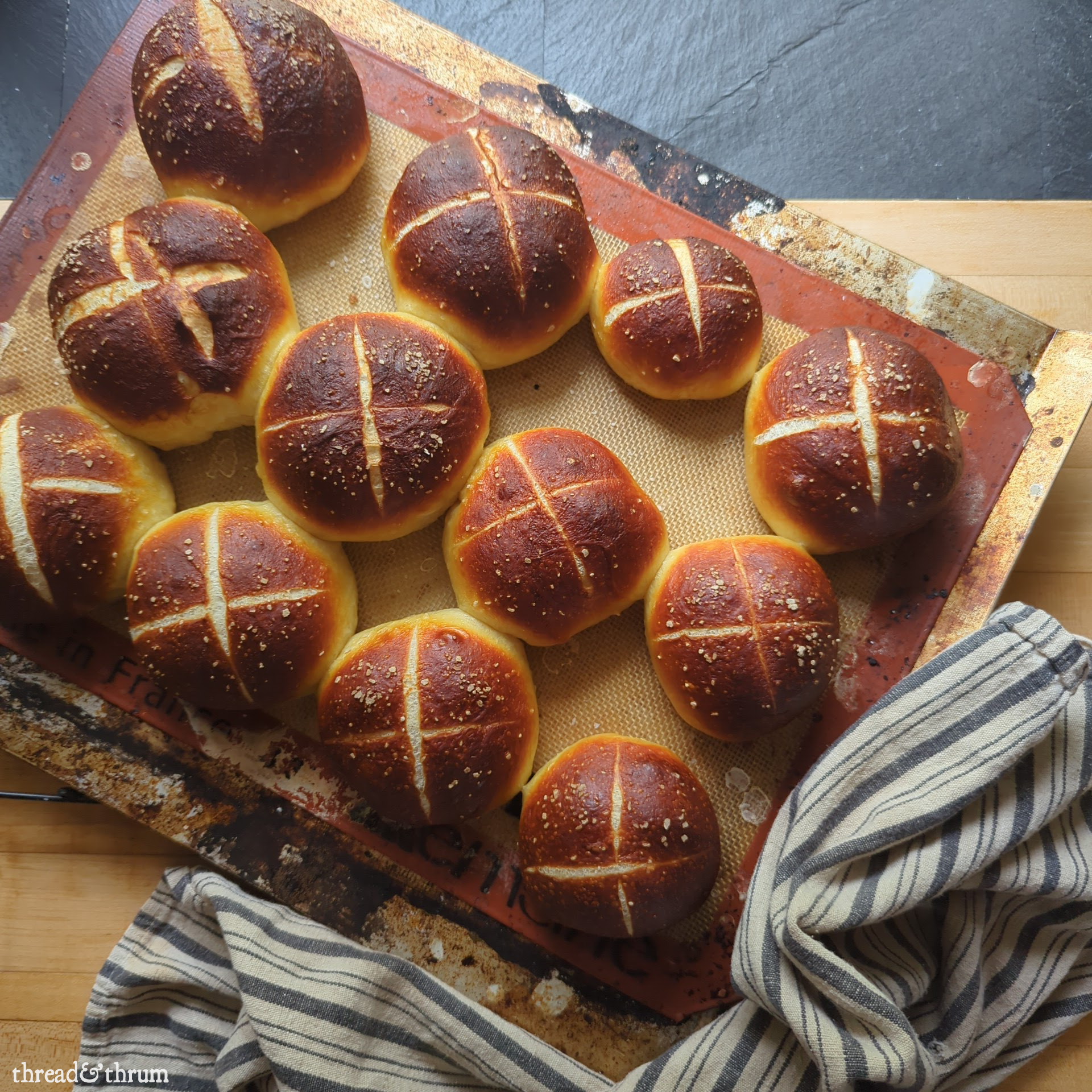 Dark coppery brown soft pretzel rolls are scored with an X on the top and are lightly salted. They sit on a silicone baking mat on a cookie sheet, which all sits on a butcher block counter